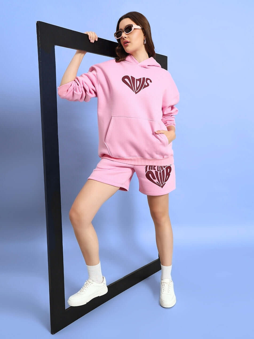 Woman modeling WOMEN LOVERS CLUB FLEECE HOODIE PINK, styled with shorts and white sneakers, standing confidently in a frame.