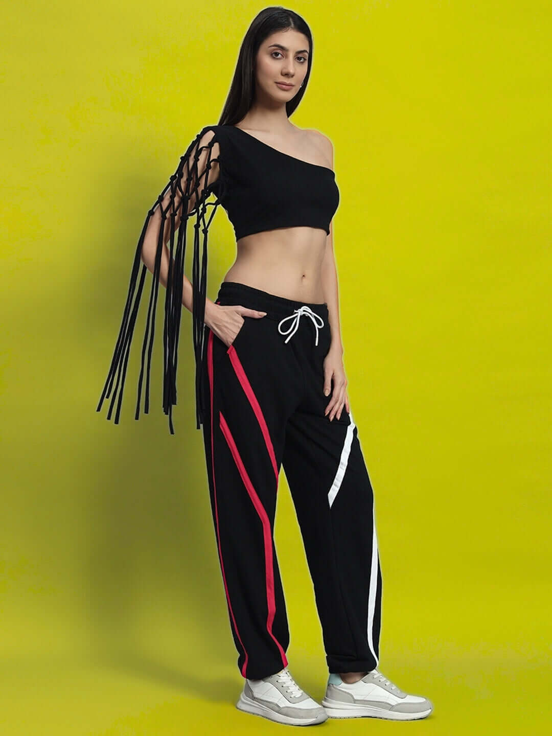 Woman wearing a black macrame crop top with fringe sleeve detail, paired with track pants, standing against a yellow backdrop.