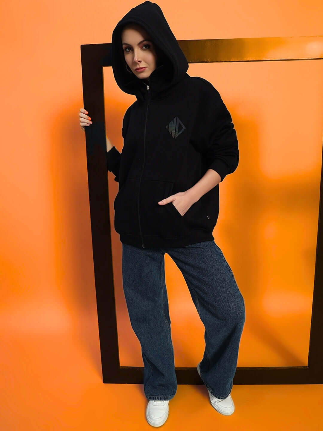 Woman wearing Marksman oversized black fleece hoodie and jeans standing in orange room, posing with hands in pockets