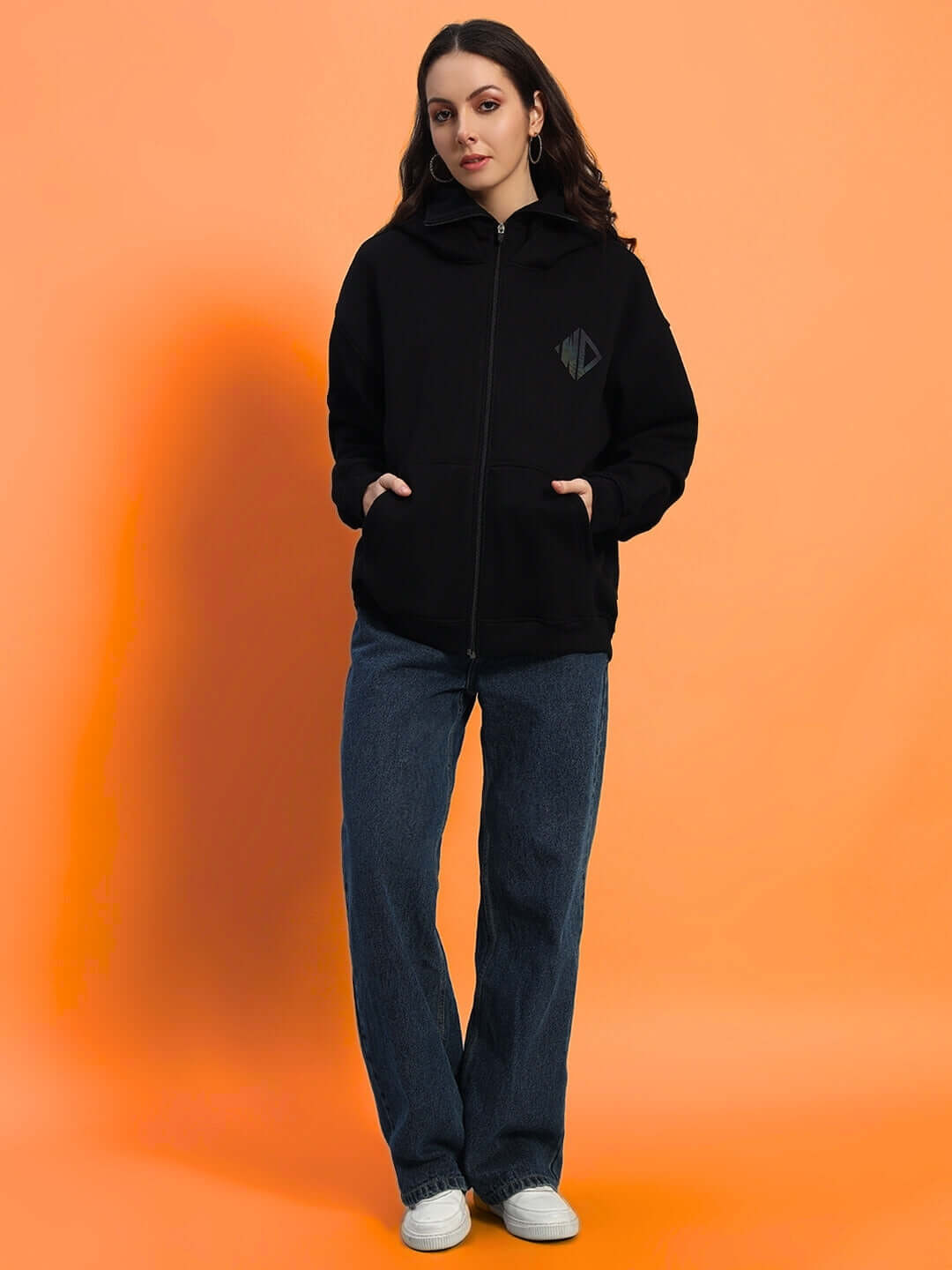 Woman wearing a black oversized fleece hoodie with logo, styled with jeans against an orange background.