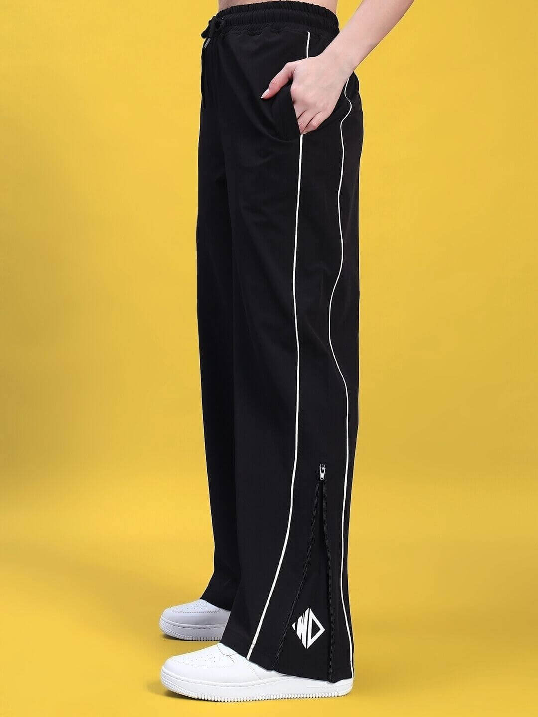 Side view of a woman wearing black joggers with white piping, styled with white sneakers, against a yellow background.