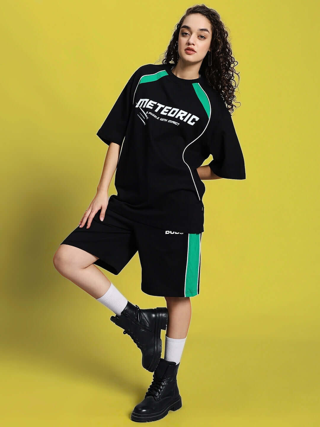 Woman wearing Women's Meteoric Regular Fit Shorts in Black with logo and green accents, styled with black boots on a yellow background.