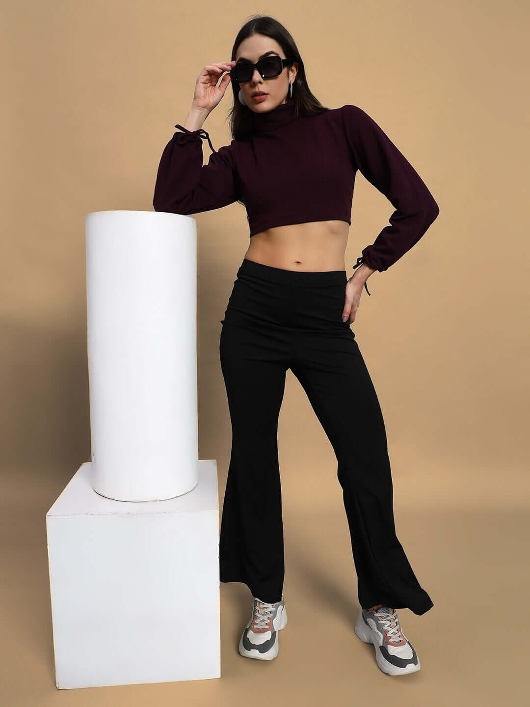 Woman wearing a wine-colored ribbed crop top made of 100% Lycra, styled with black pants and sneakers, posing against a beige background.