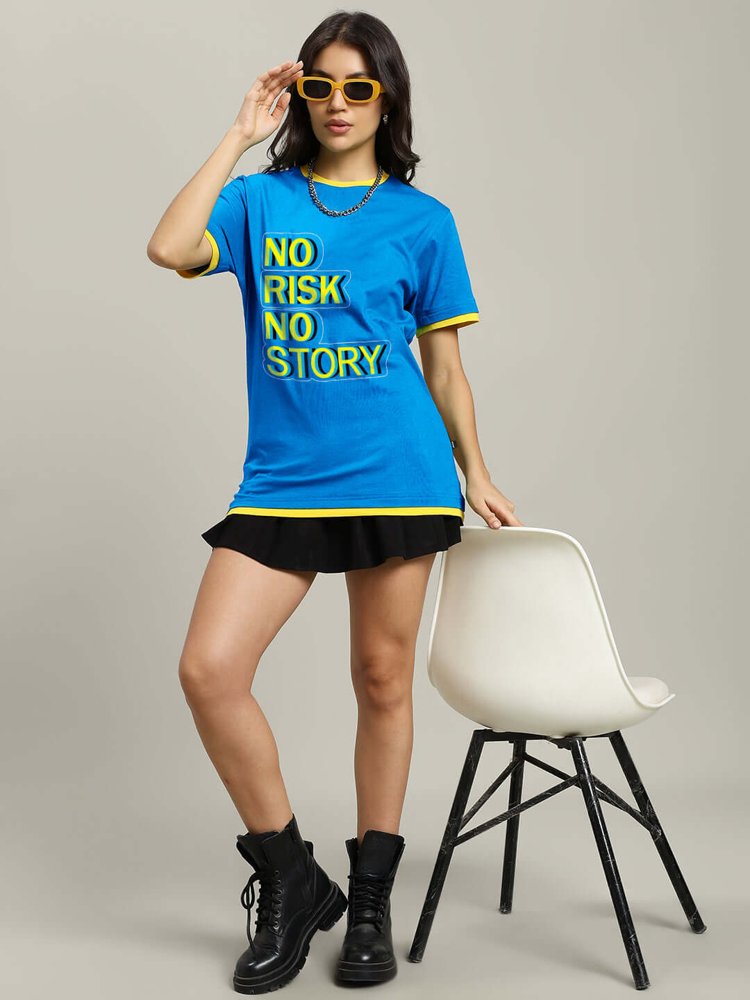 Woman in blue 'No Risk No Story' t-shirt with boots and skirt, standing by a chair, promoting women's fashionable casual wear.