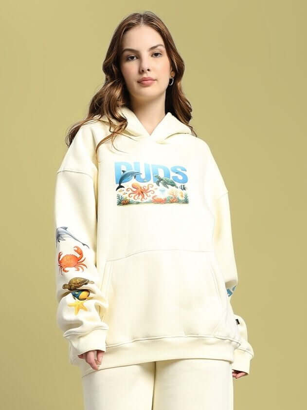 Woman wearing WOMEN OCEAN BUDDIES OVERSIZED FLEECE HOODIE OFF-WHITE with ocean-themed graphics, emphasizing cozy and stylish loungewear.