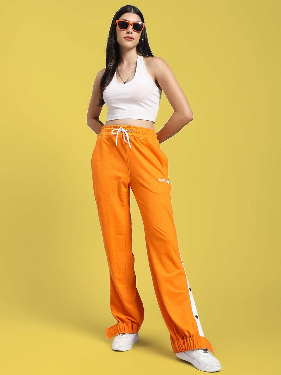 Model wearing WOMENS MILLER RELAXED FIT JOGGERS ORANGE with white top and sunglasses, showcasing vibrant casual style on yellow background.