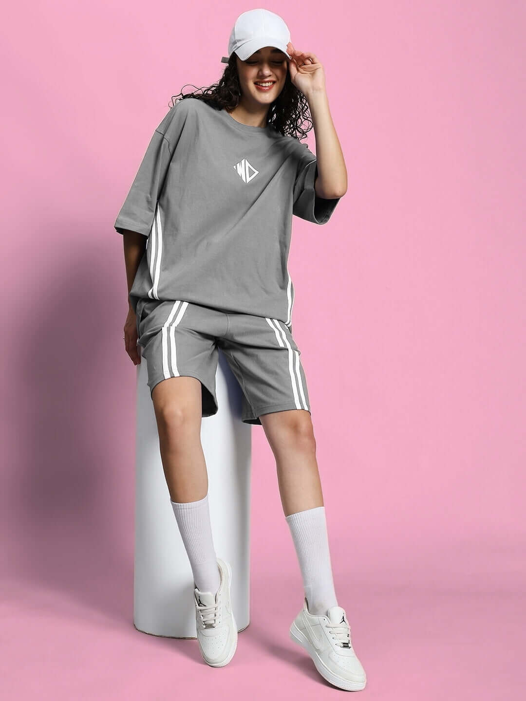 Woman wearing WOMEN'S OUTLOOK SUMMER CO-ORD SHORTS SET GREY with casual sneakers and cap, posing stylishly against a pink backdrop.