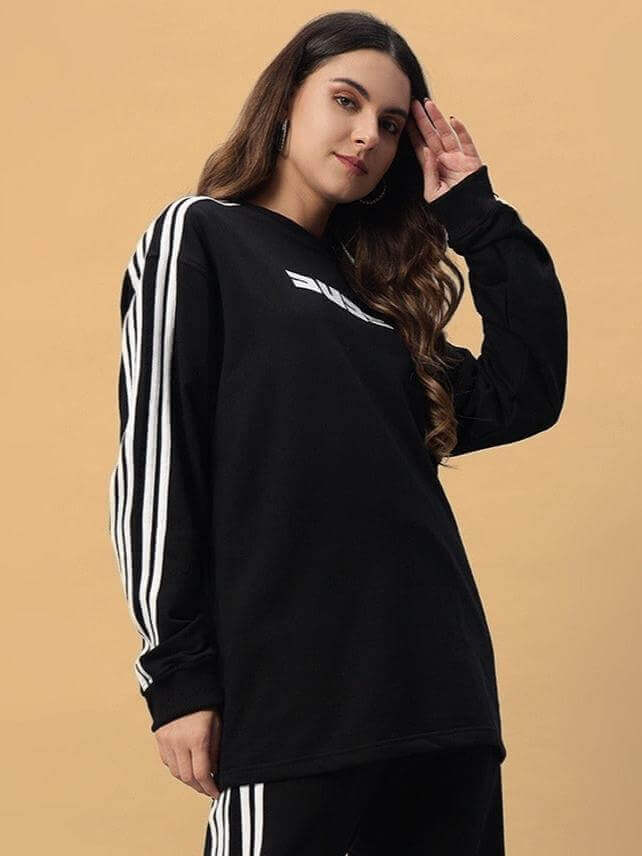 Young woman wearing an oversized black women's parka sweatshirt with white stripe sleeves, made from premium cotton blend, standing against a tan background.