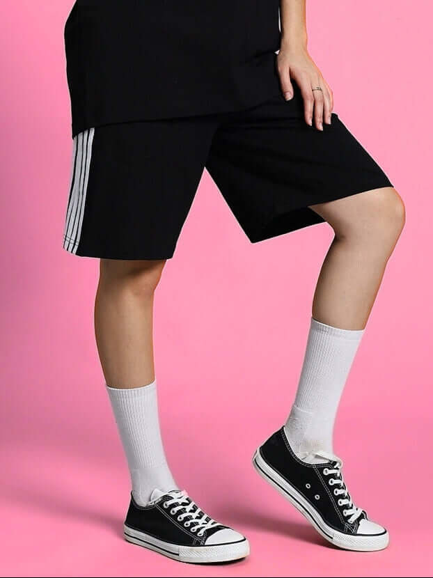 Woman wearing WOMEN'S PARKA REGULAR FIT SHORTS BLACK paired with white socks and sneakers against a pink background.