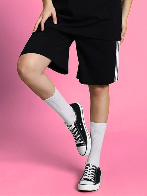 Woman wearing black regular fit shorts with white socks and black sneakers against a pink background.