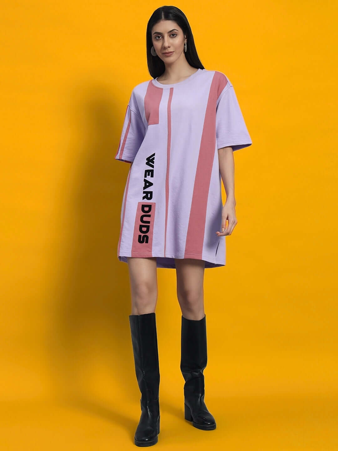 Woman wearing a lilac dress with vertical pink stripes and black boots standing against an orange background.