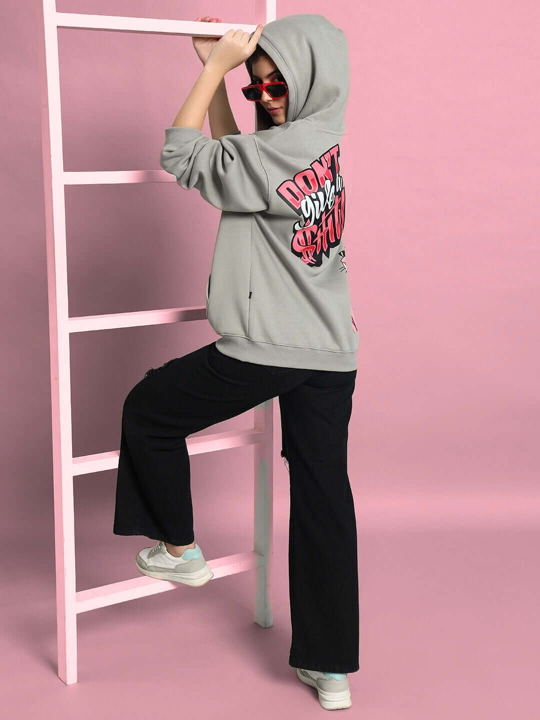 Model wearing WOMEN PINK PANTHER OVERSIZED HOODIE GREY with graphic text, paired with black pants and pink backdrop.