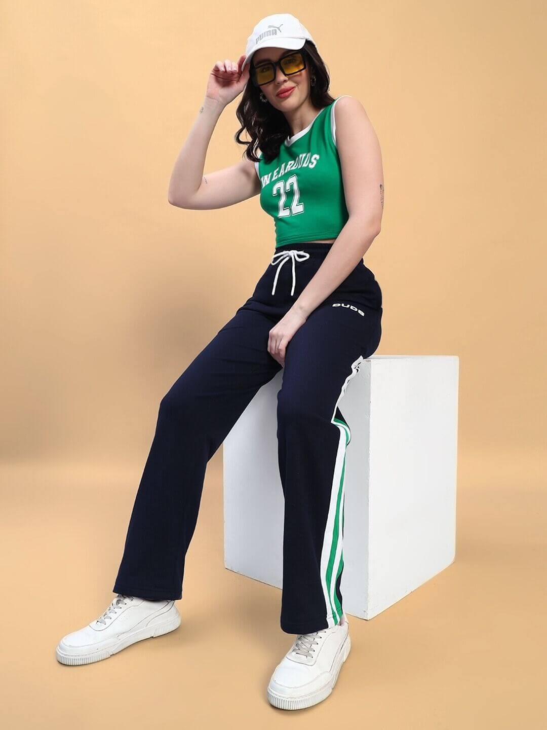 Woman wearing WOMEN'S PLAYER CO-ORD SET GREEN NAVY, seated on a white cube against a beige background, showcasing sporty fashion.