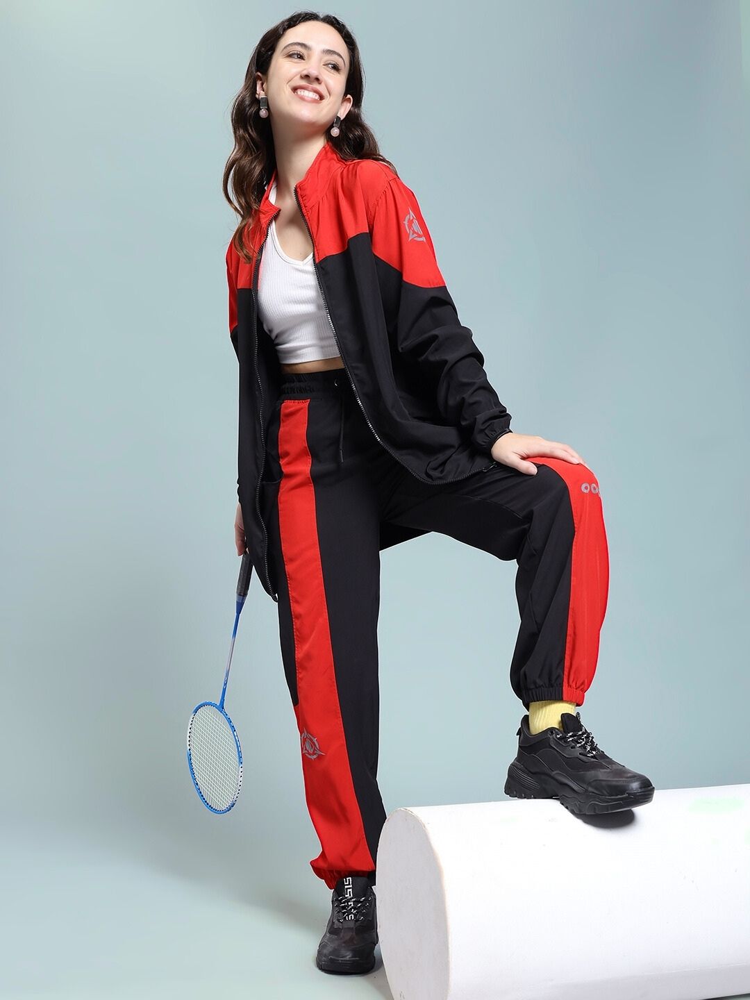 Woman wearing WOMEN'S POWER BLOCK RELAXED FIT JACKET BLACK RED holding a badminton racket, striking a sporty pose.