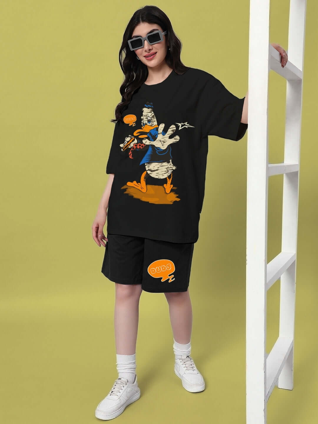 Woman wearing black Quack Off summer shorts and graphic tee, stylish sunglasses, standing by ladder against light green background.