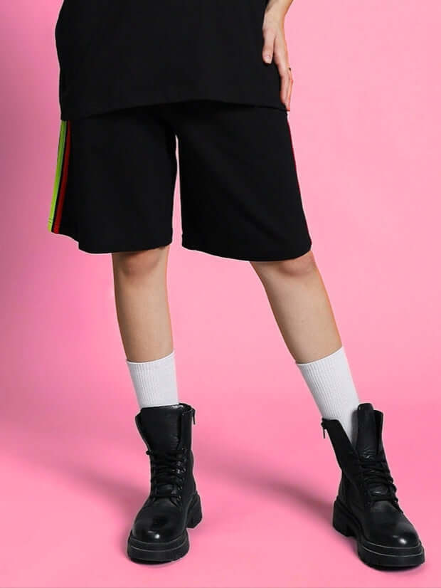 Model wearing Women's Rainbow Regular Fit Shorts in black with rainbow stripes, black boots, and white socks, standing against a pink background.