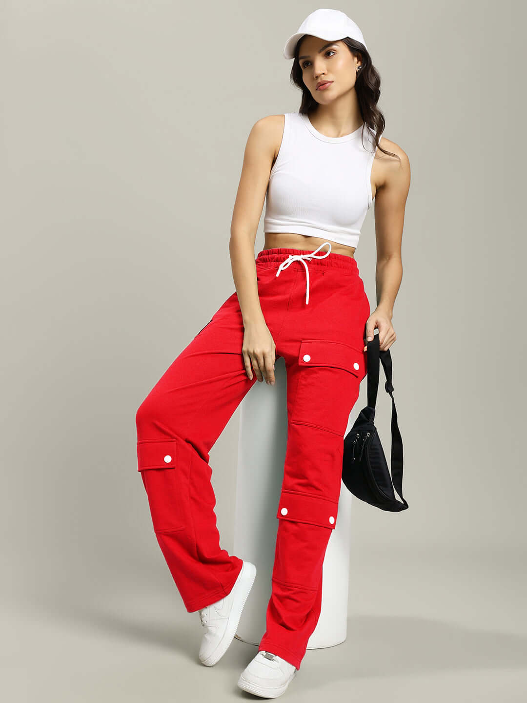 Woman wearing WOMENS AWESOME FLAP POCKET BAGGY JOGGER RED with white top and cap, holding a black bag, showcasing casual streetwear style.