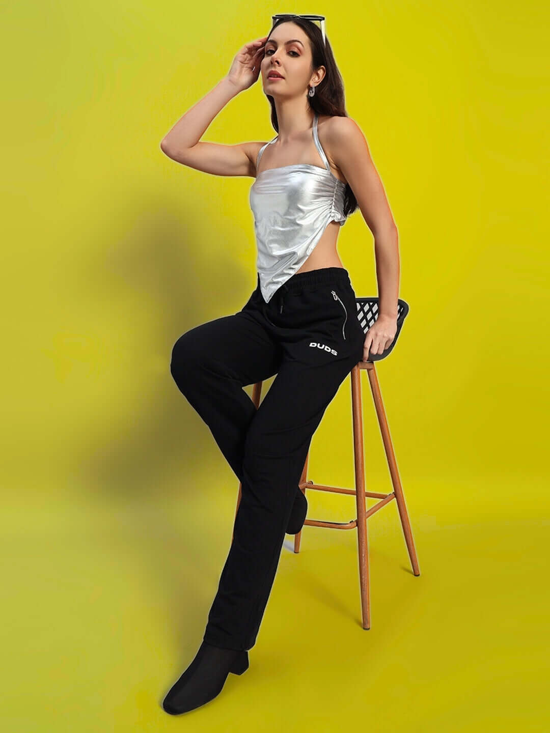 Woman wearing WOMEN'S REGIONAL CROP TOP SILVER, metallic silver crop top with modern chic design, seated on a stool against yellow background.