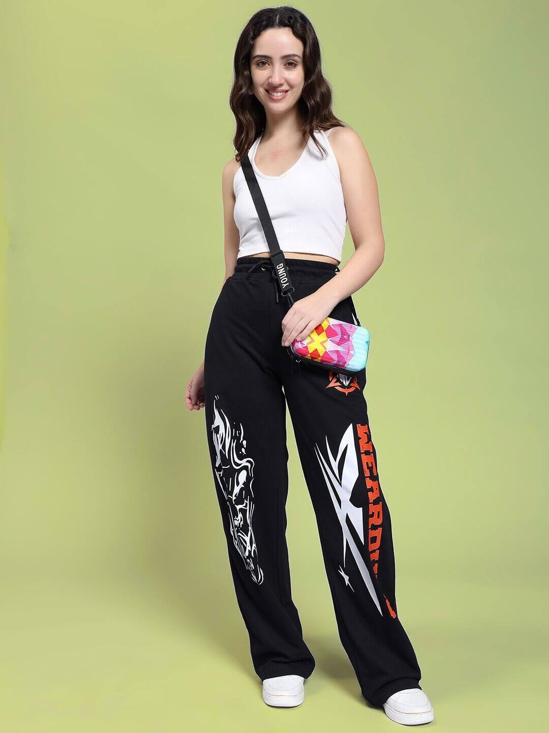 Woman wearing WOMEN RIFT RELAXED FIT JOGGER IN BLACK with graphic print, paired with a white tank top and crossbody bag, standing in studio.