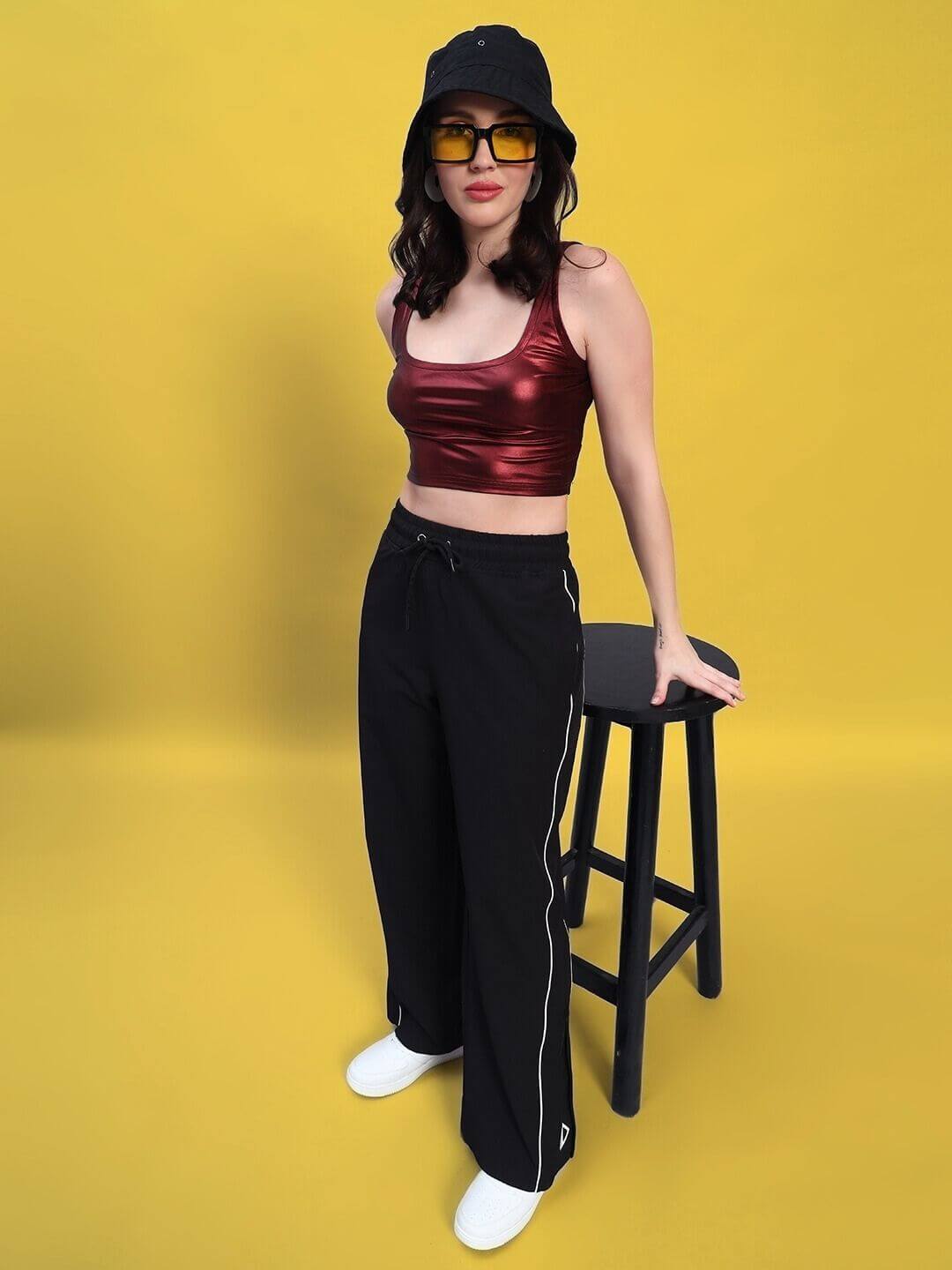 Model wearing women's relaxed-fit joggers in black, paired with a red top, yellow sunglasses, and a black hat, sitting on a stool.