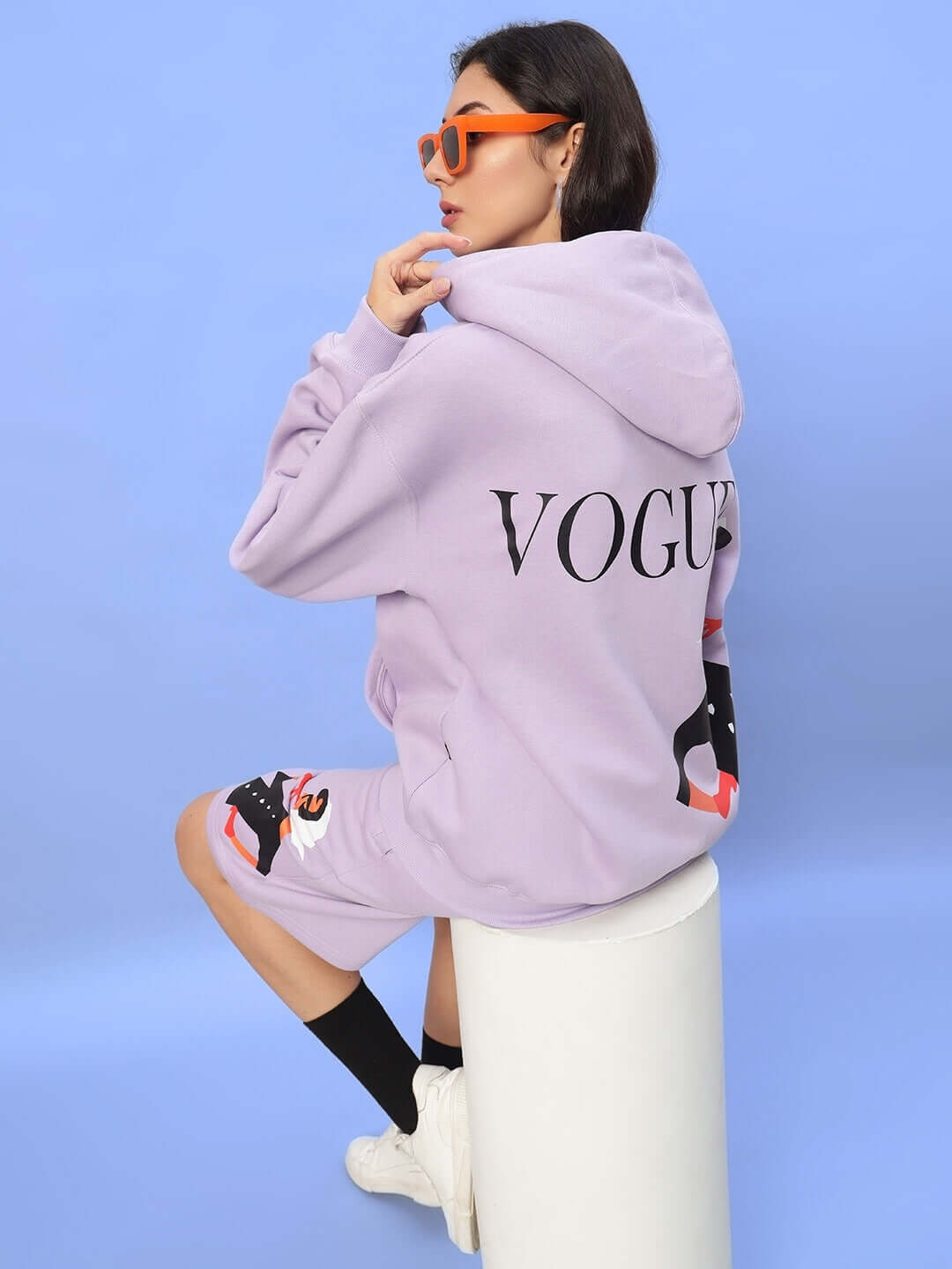 Woman wearing WOMEN RITZY FLEECE CO-ORD LILAC, featuring a lilac hoodie and shorts, sitting on stool with orange sunglasses.