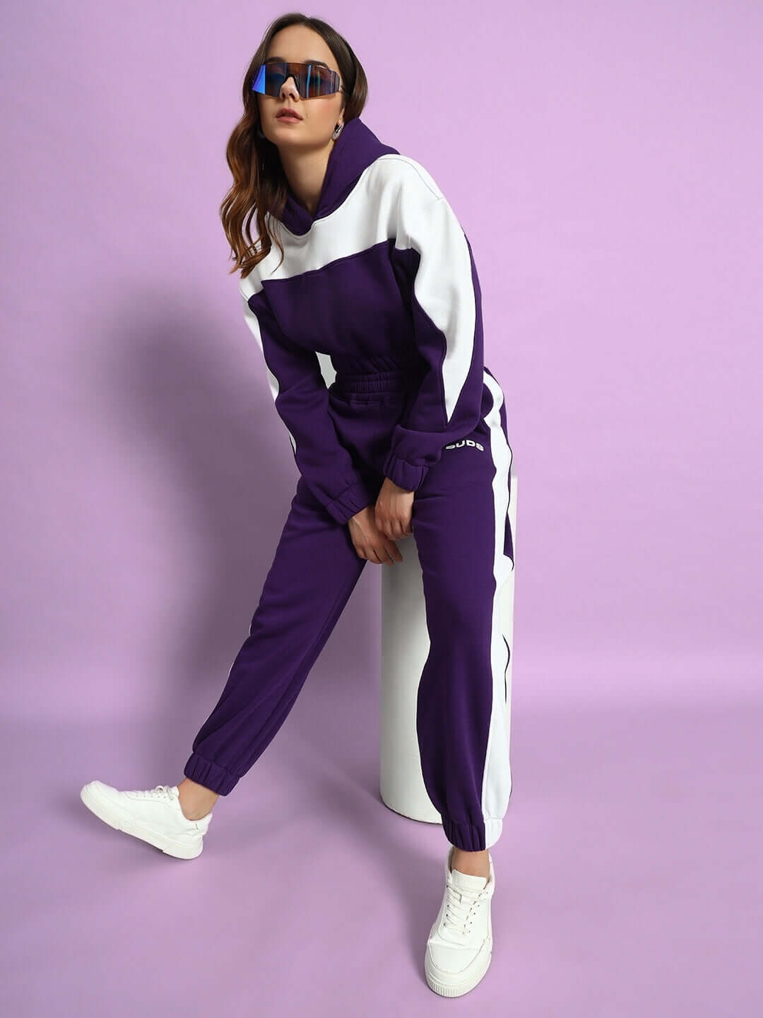 Model wearing WOMEN RIZZLER FLEECE OVERSIZED CROP HOODIES PURPLE with colorblock accents and white sneakers, sitting on stool.