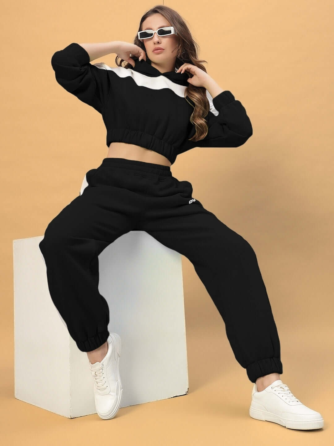 Woman wearing WOMEN RIZZLER OVERSIZED CROP HOODIES BLACK, sitting stylishly on a white block with sunglasses.