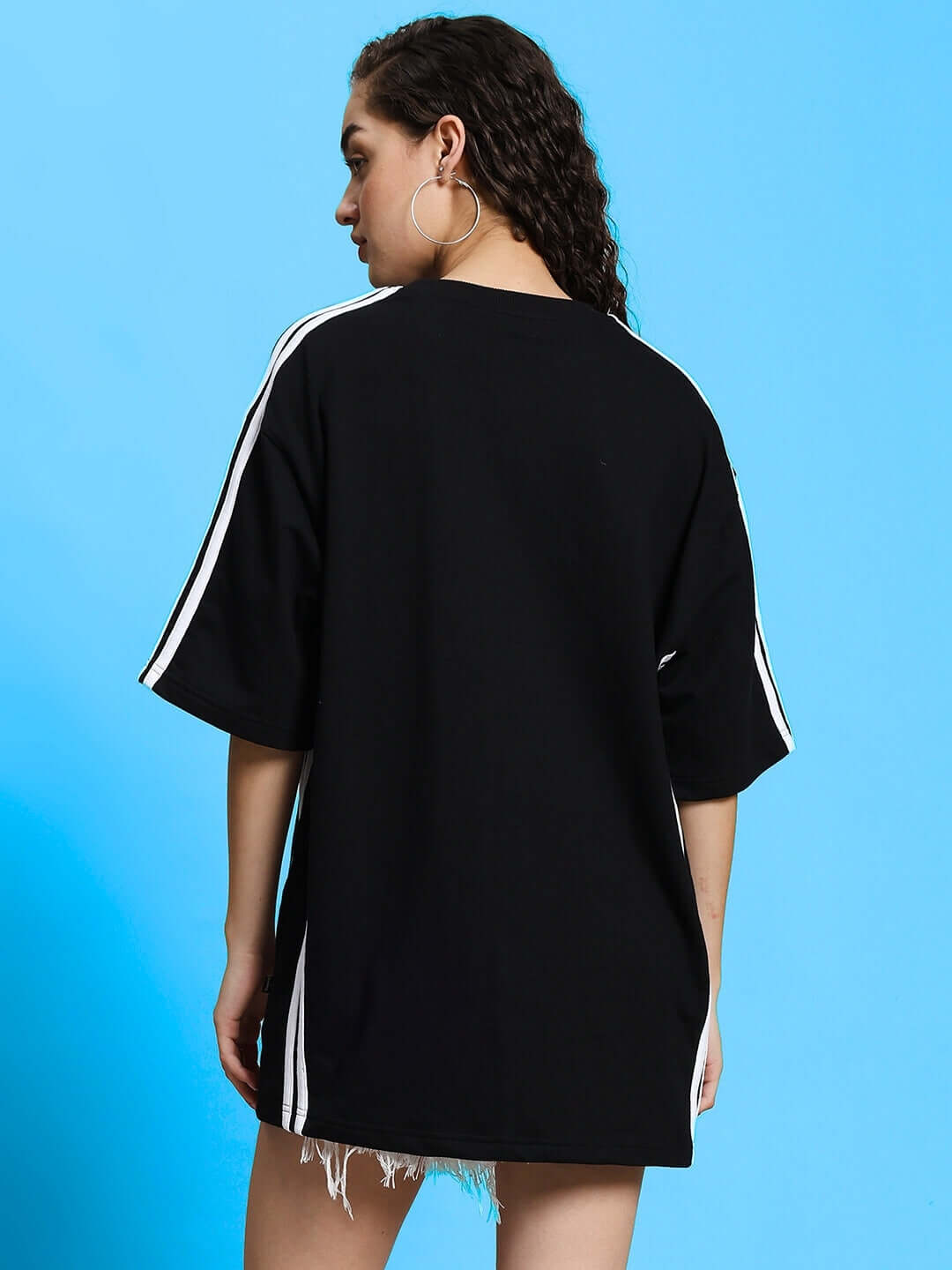 Back view of a woman wearing a black Roddy oversized classic T-shirt with white stripes and distressed shorts on a blue background.