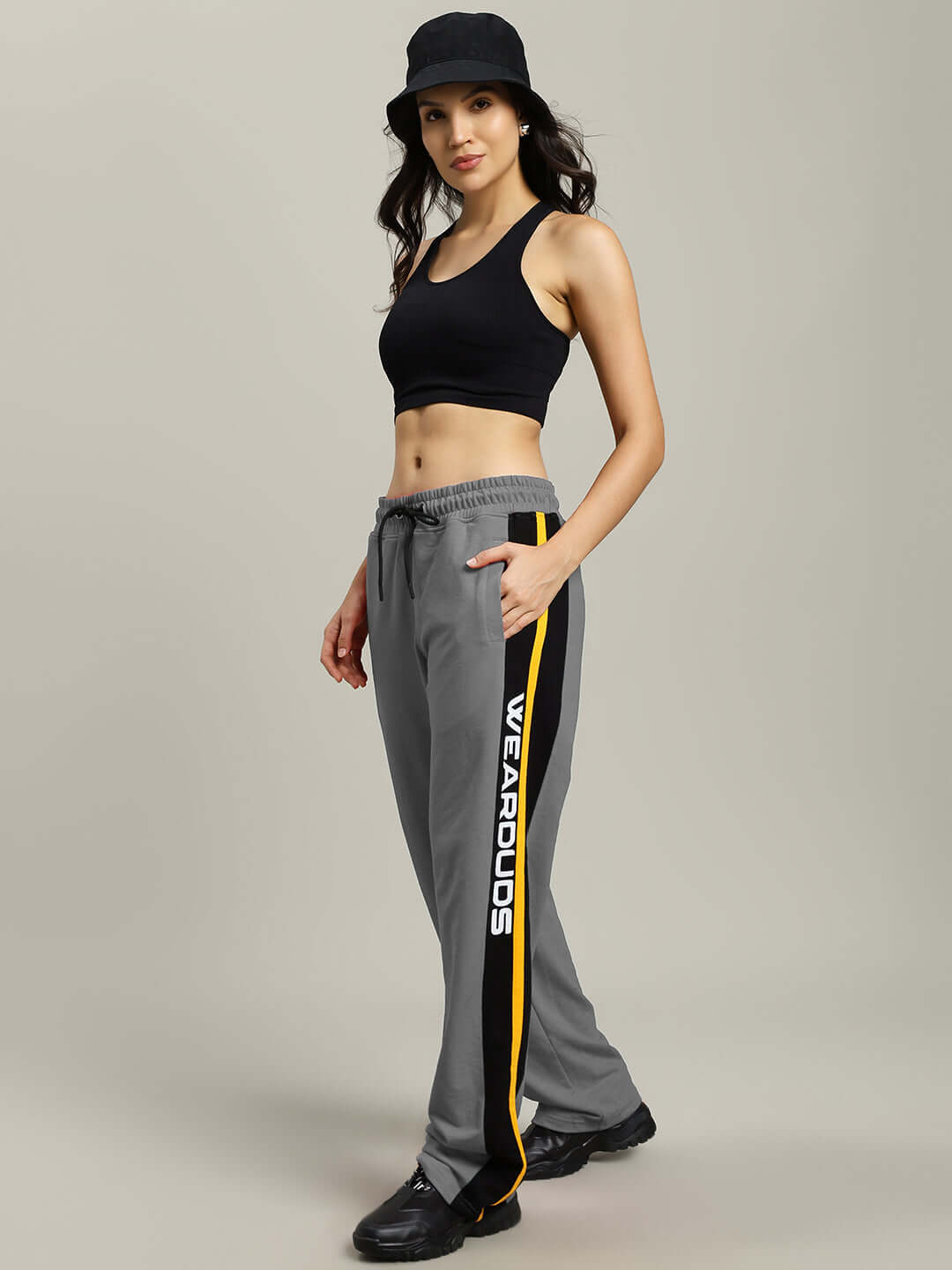 Woman wearing grey relaxed-fit joggers with 'WEAR DUDS' branding and a black top and hat.