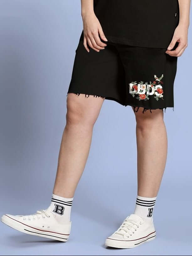 Woman wearing WOMEN ROSE CHARM RELAXED FIT SHORTS BLACK with floral design, paired with white sneakers against a blue background.