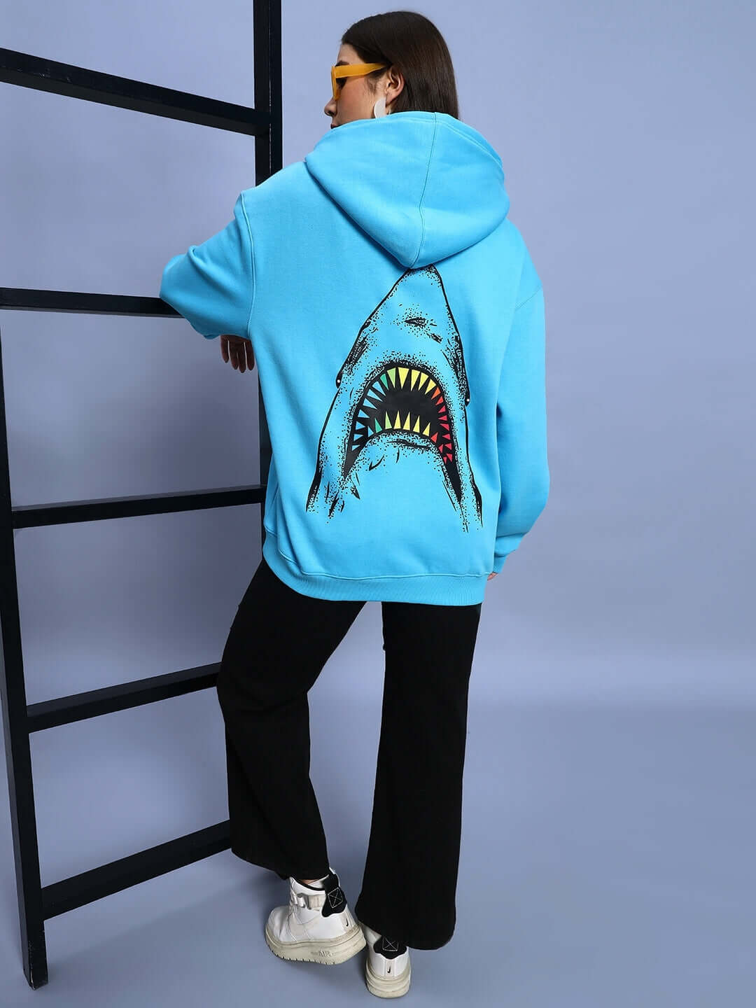 Woman wearing WOMEN SHARKWAVE FLEECE OVERSIZED HOODIE LIGHT BLUE with shark graphic, standing by ladder, showcasing streetwear style.