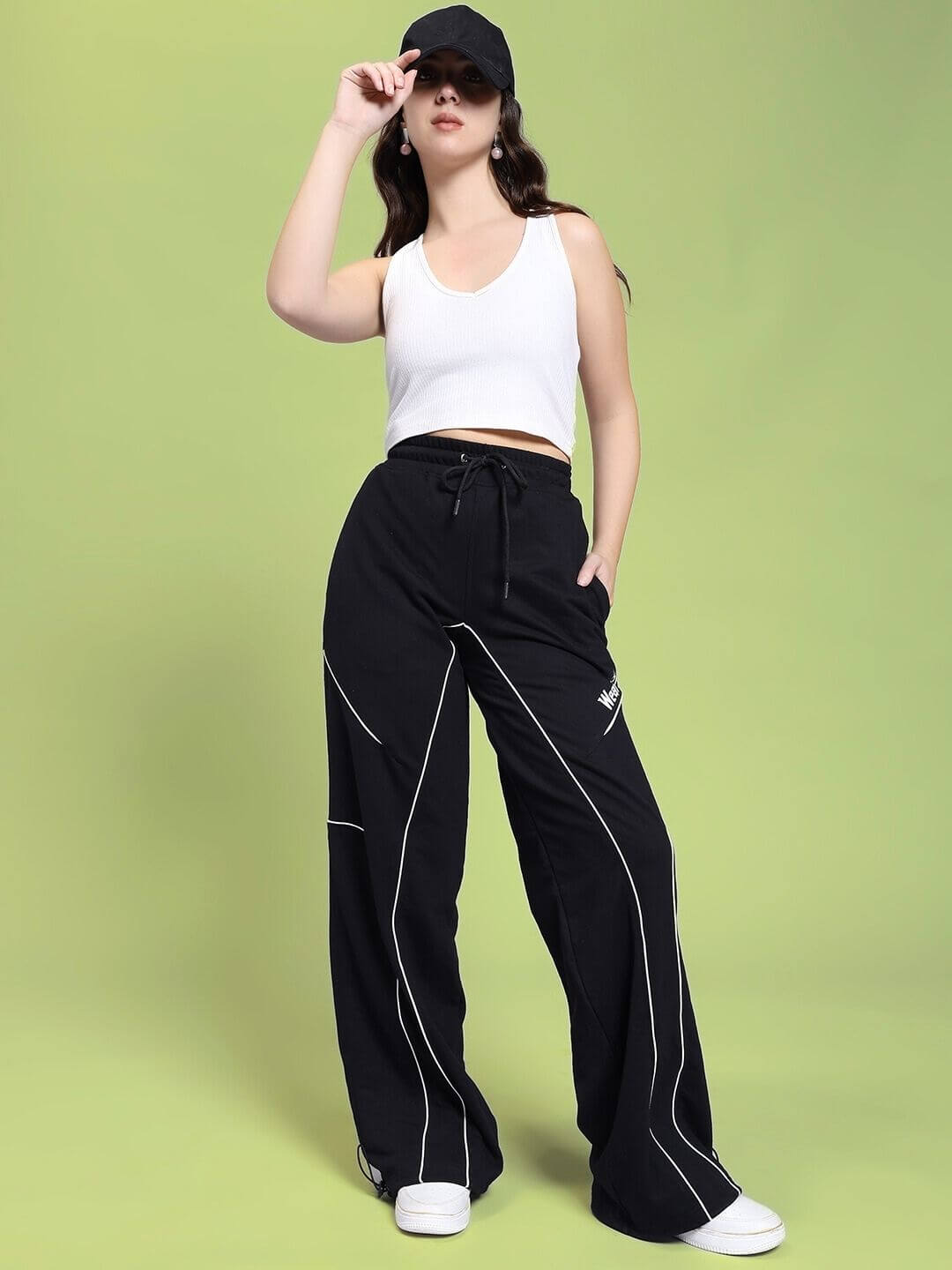Model wearing WOMEN SIGNATURE PIPING JOGGERS BLACK with contrast piping, styled with a white tank top and black cap in a casual pose.