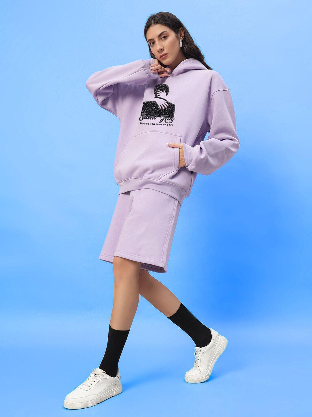 Woman wearing WOMENS SILENT HUG PRINTED FLEECE CO-ORD LILAC set with hoodie and shorts, showcasing cozy street style on blue background.