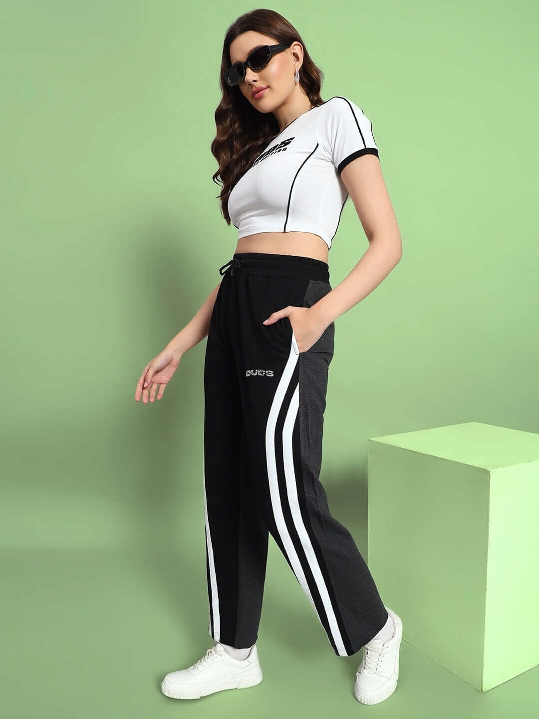 Woman wearing black and grey skater Korean baggy joggers with white stripes and a white crop top, standing against a green background.