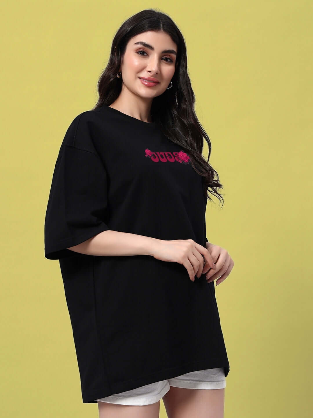 Woman wearing a black oversized t-shirt with pink skull and roses design, styled with white shorts against a yellow background.