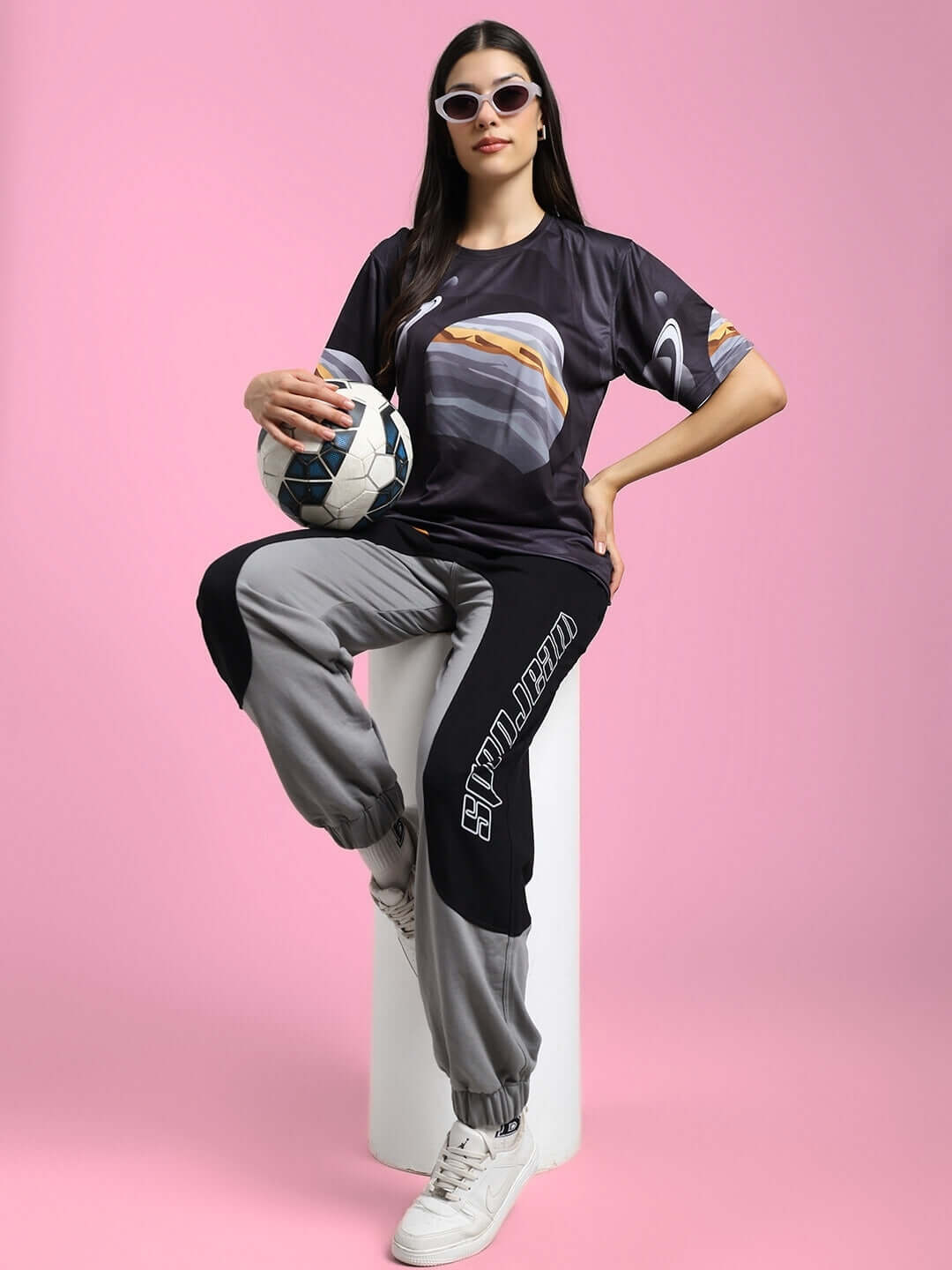Woman in black space fantasy printed t-shirt, holding soccer ball, seated on pink background, showcasing sporty fashion.