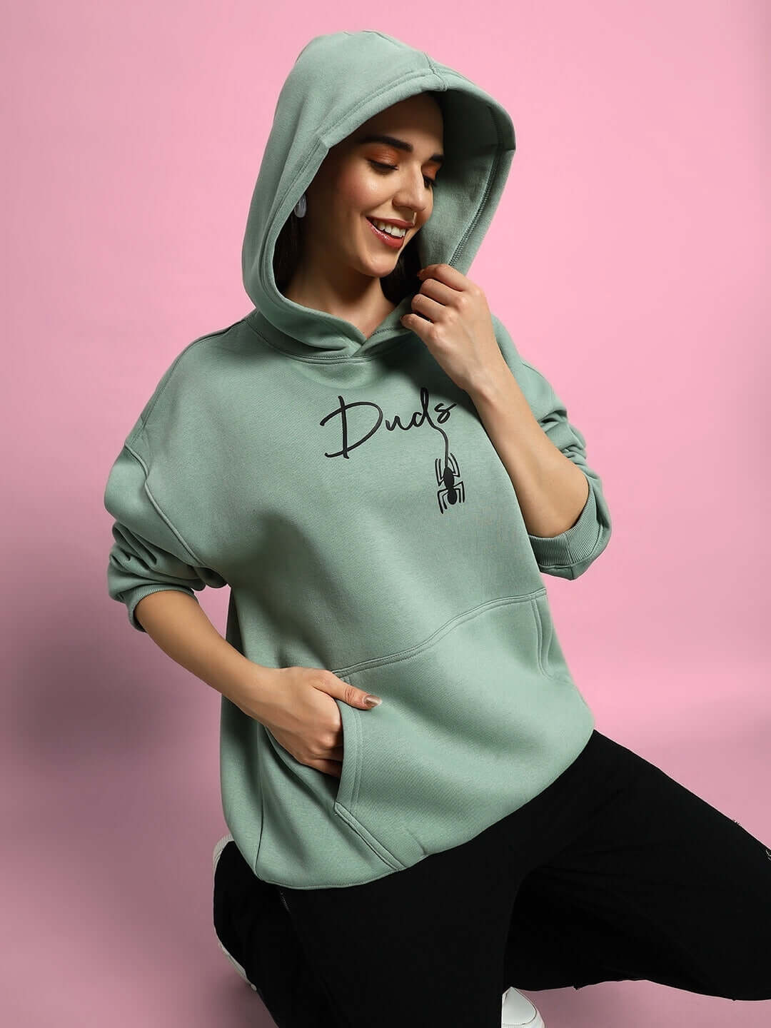 Woman wearing WOMEN SPIDERMAN OVERSIZED FLEECE HOODIE JADE GREEN, showcasing comfort and style on a pink backdrop.