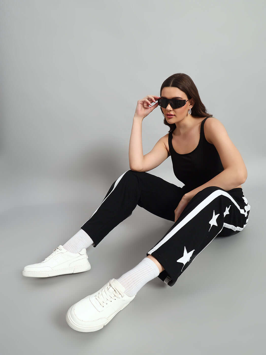 Woman sitting in black joggers with star pattern, wearing sunglasses and white sneakers against a neutral background.