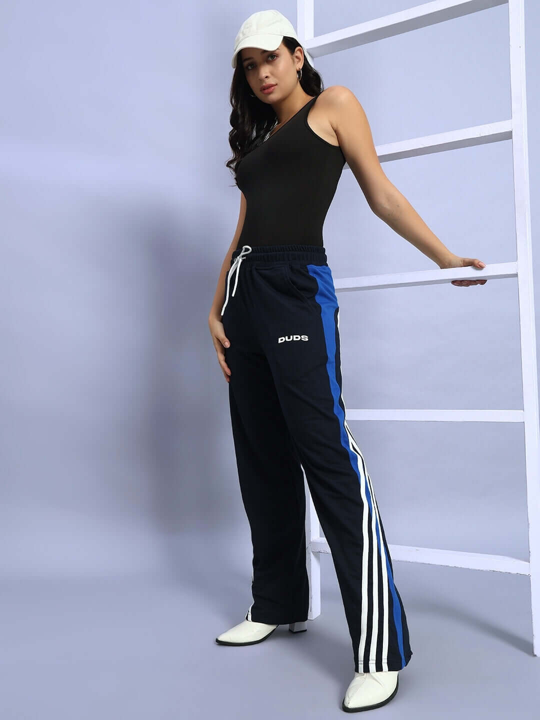 Model wearing Women's Tangle Relaxed-Fit Joggers in Navy Blue with white stripes, paired with a black top and white cap, leaning on ladder.