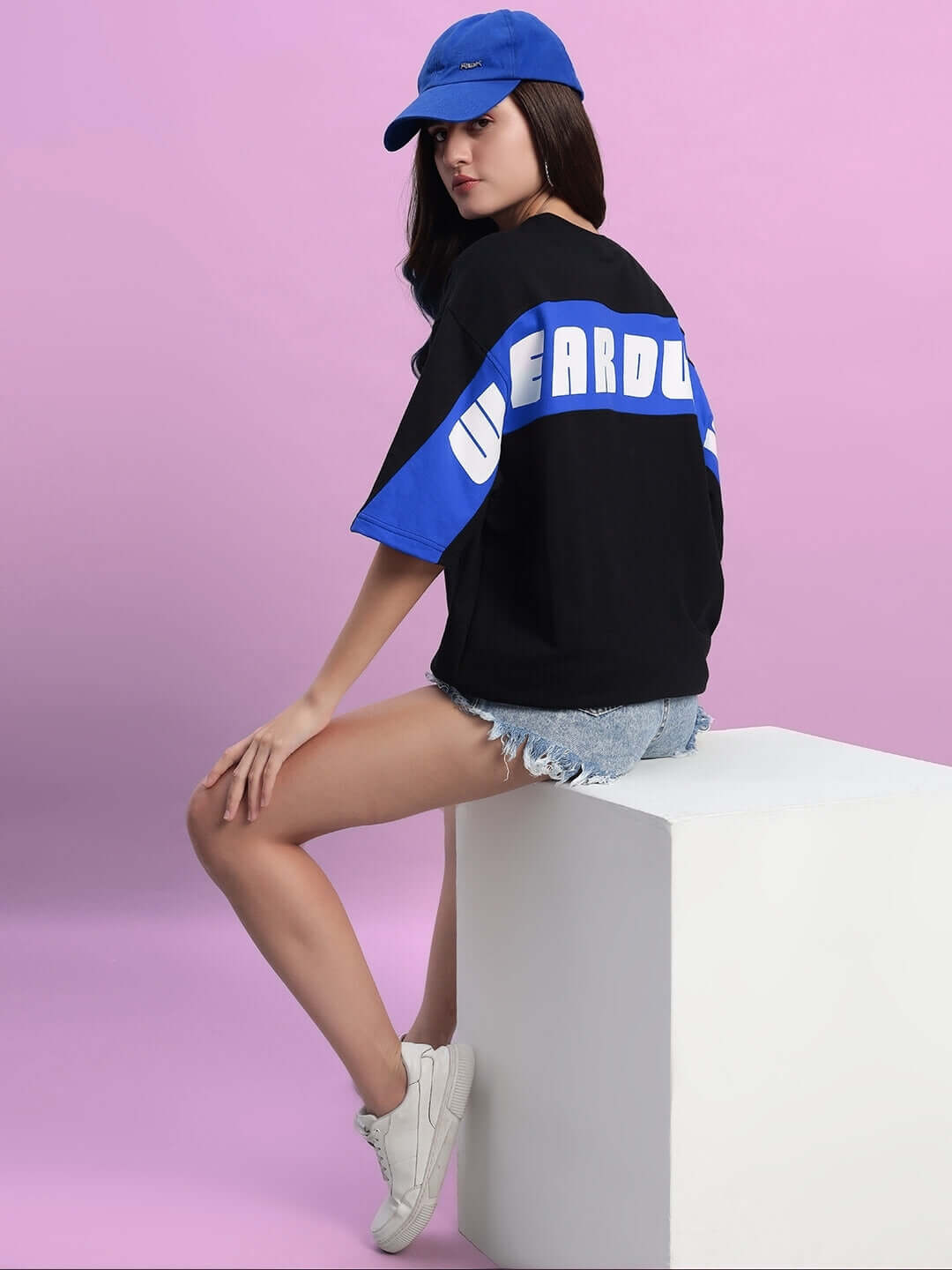 Woman wearing oversized black-blue t-shirt with text and cap, sitting on a white cube against a pink background.