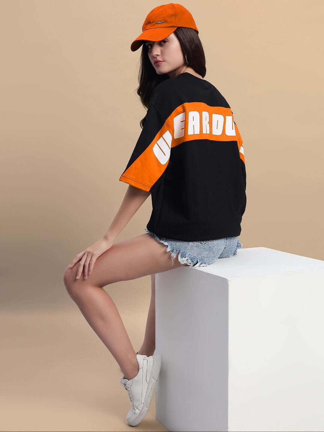 Model wearing Women's Titan oversized black-orange t-shirt with text, paired with shorts and cap, seated on a white block, side view.