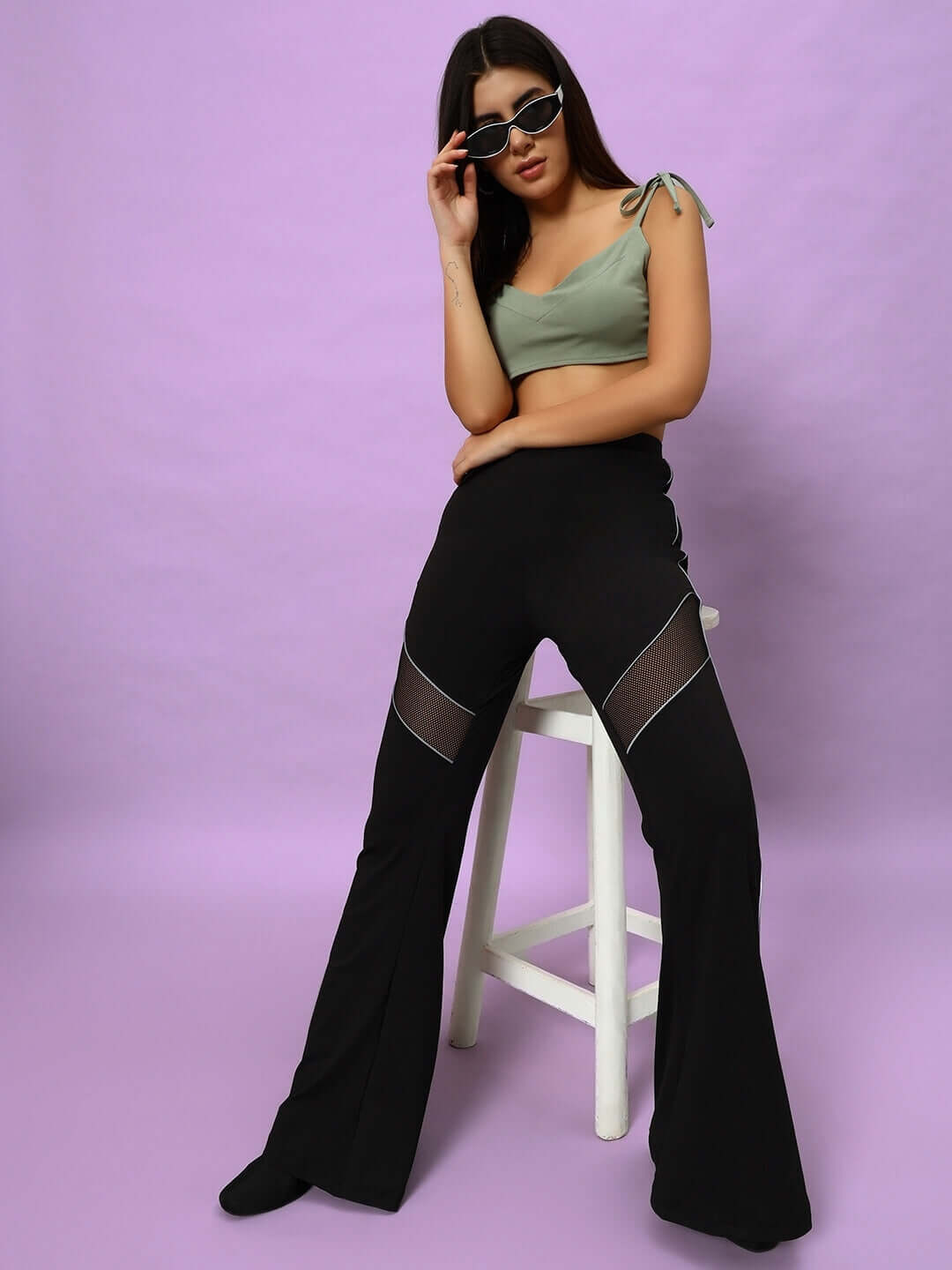 Model wearing black women's Victoria boot cut reflector pants, sitting on a stool against a purple background, styled with sunglasses.