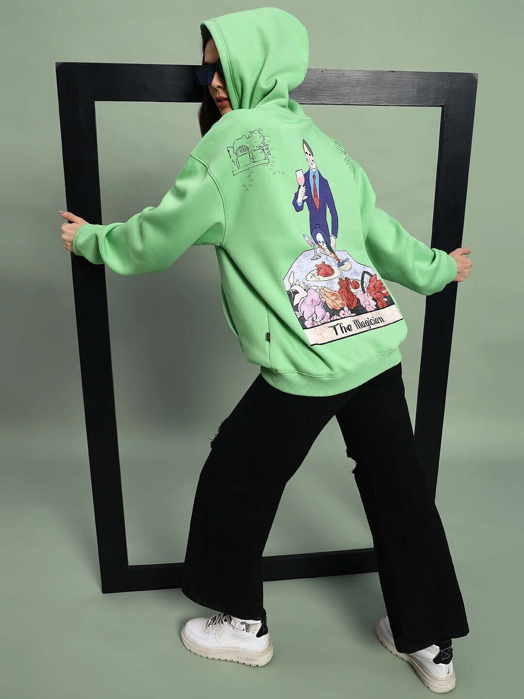 Woman wearing WOMEN VOODO FLEECE OVERSIZED HOODIE GREEN with graphic art on back in studio.