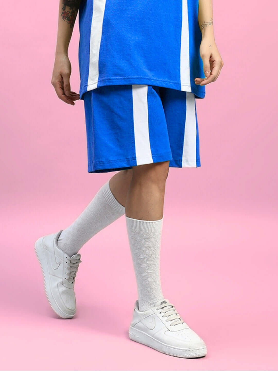 WOMENS WIDELINE REGULAR FIT SHORTS BLUE showcased with a casual outfit including white sneakers and socks against a pink background.