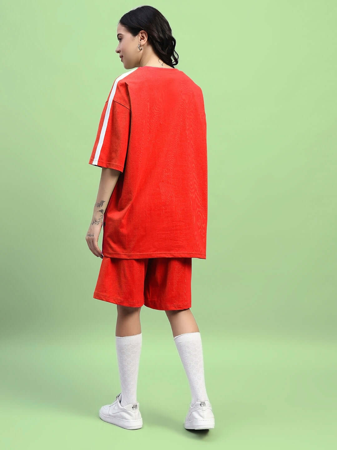 Woman wearing WOMENS WIDELINE REGULAR FIT SHORTS RED with matching oversized shirt, standing against a green backdrop.