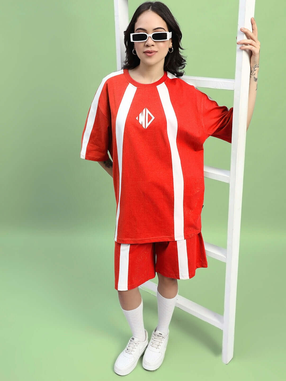 Woman wearing red women's wideline regular fit shorts and matching top, standing next to a ladder, against a green background.