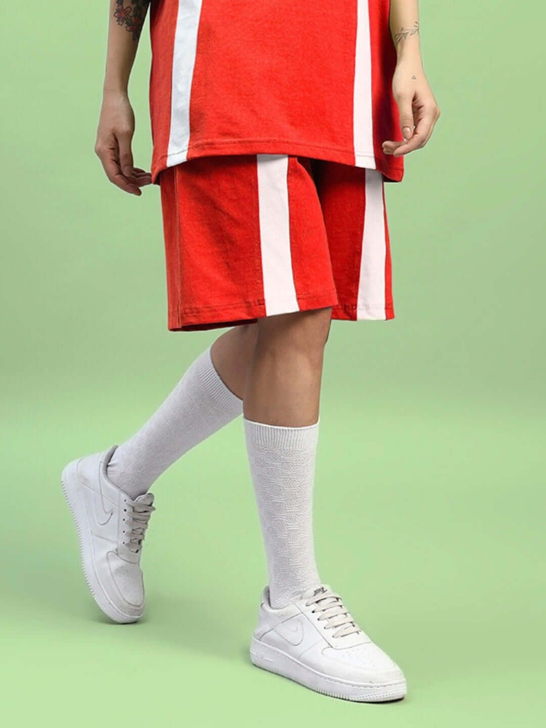Women's red wideline regular fit shorts with white stripes, styled with white sneakers and knee-high socks on a green background.