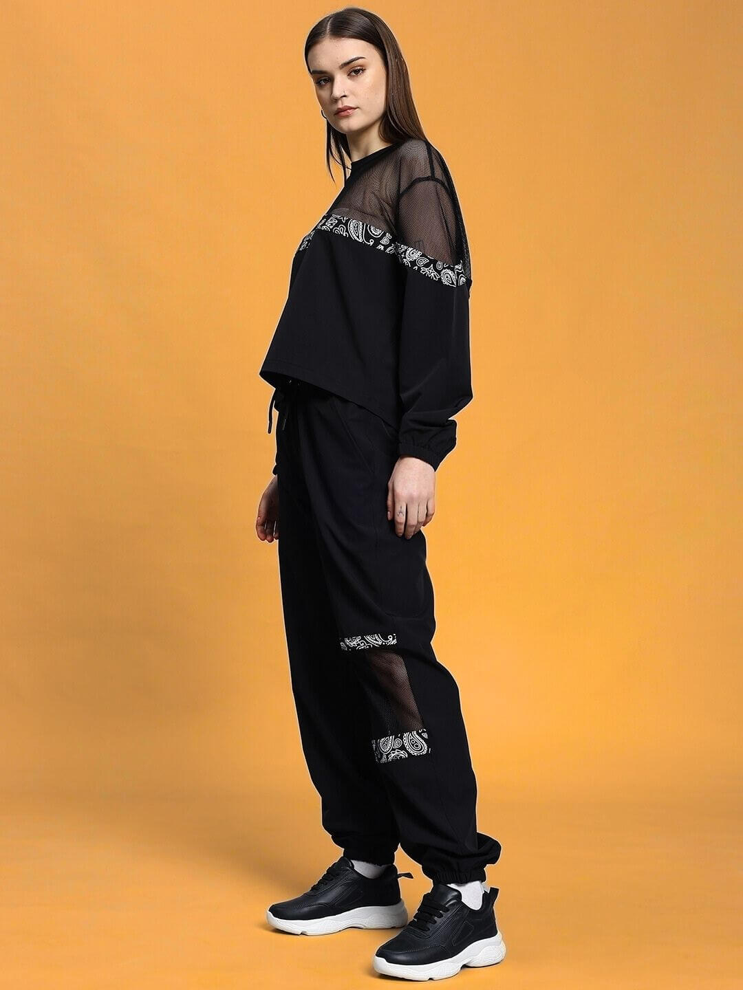 Model in women's black relaxed fit co-ord set with sheer details, elastic waistband, and sneakers, posed against a yellow background.