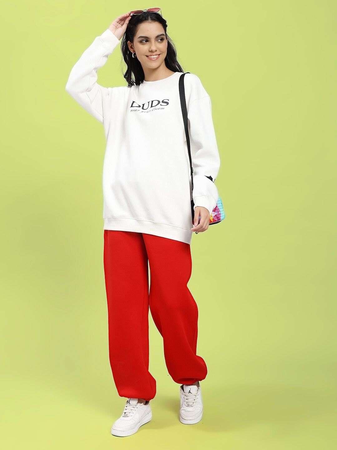 Woman wearing WOMEN'S ZENO RELAXED FIT FLEECE JOGGER RED with white sweatshirt and sneakers on a green background.