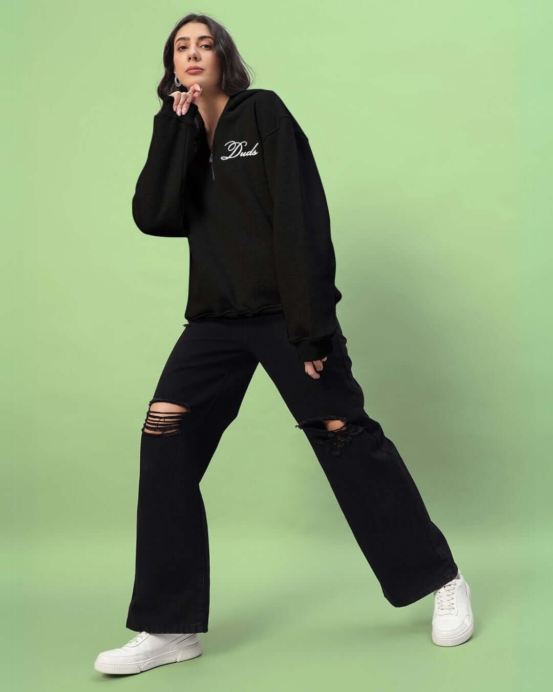 Woman wearing Amarillo black fleece hoodie with ripped jeans and white sneakers, standing against a light green background.